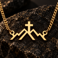 A gold-toned chain necklace with a pendant shaped like stylized mountains and a cross, displayed on a dark textured rock against a dark background.
