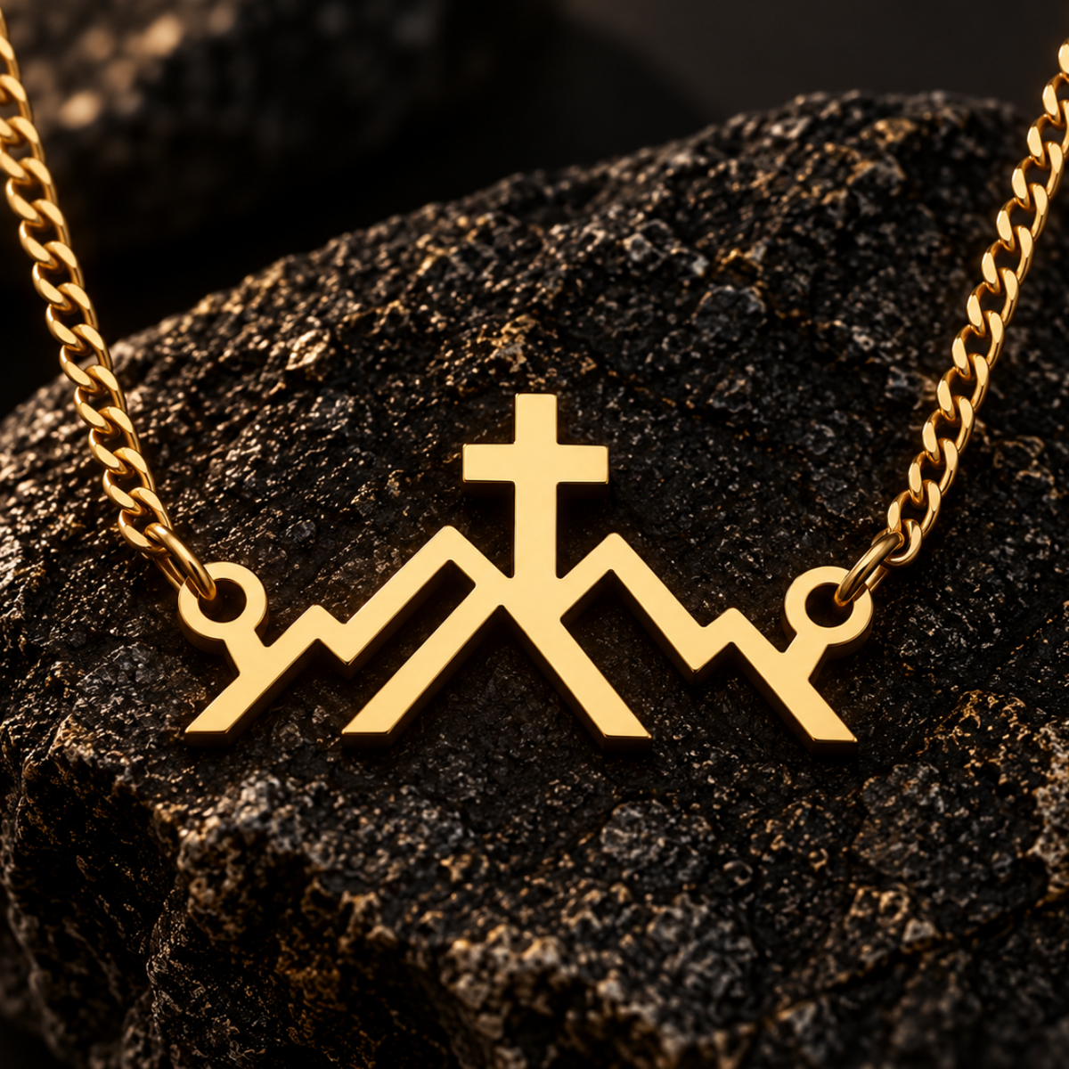A gold-toned chain necklace with a pendant shaped like stylized mountains and a cross, displayed on a dark textured rock against a dark background.